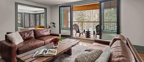 A living room with a brown leather sofa, a wooden coffee table with a book, and a view through large sliding glass doors to an outdoor deck.