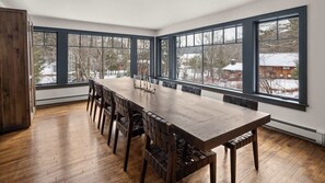 A spacious dining area with a long wooden table and chairs, large windows with dark frames, and a view of a snowy landscape.