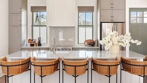 A kitchen with white cabinetry, a central island with bar stools, a stainless steel refrigerator, and a window with a view of trees. - Spacious Savannah Home | Pool, Spa & Greens (Savannah)