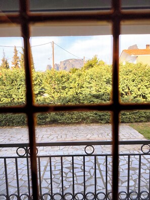 A view through a window with a metal grille, featuring a stone-paved area, green foliage, and distant buildings. - Kastraki House with a view (Kalabaka)