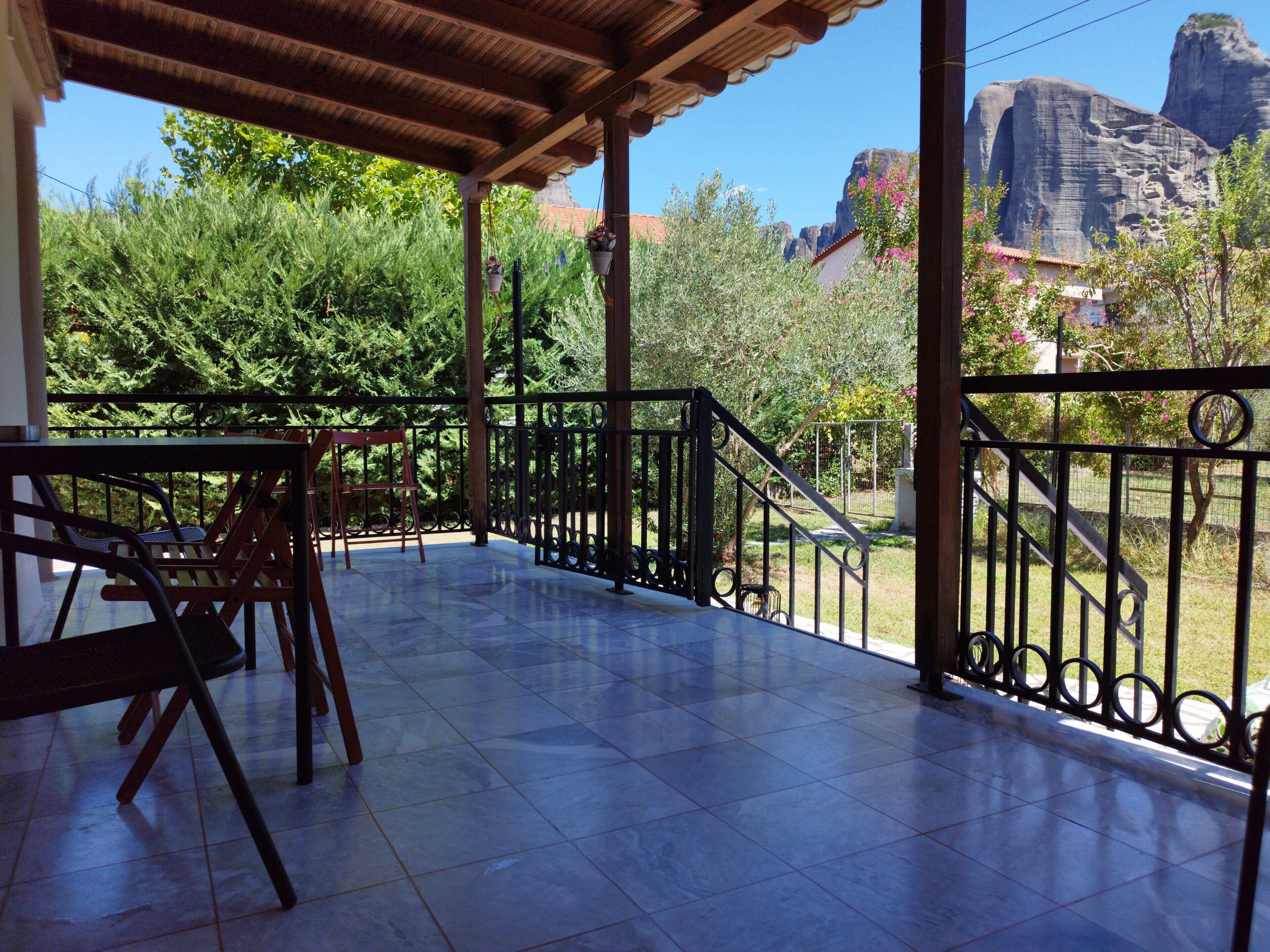 Un patio cubierto con suelo de baldosas, una mesa de madera y sillas. Se observa una barandilla metálica y, al fondo, un paisaje montañoso.