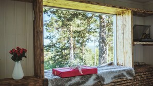 A rustic cabin interior with a wooden bench, a vase with red flowers, and a window with a view of a forest.