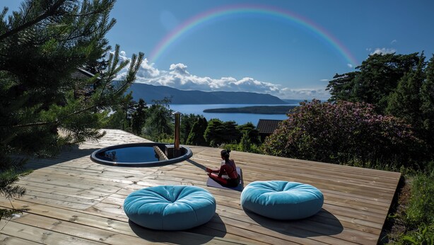 A wooden deck with two blue bean bags, a hot tub, and a view of a lake with mountains in the background.