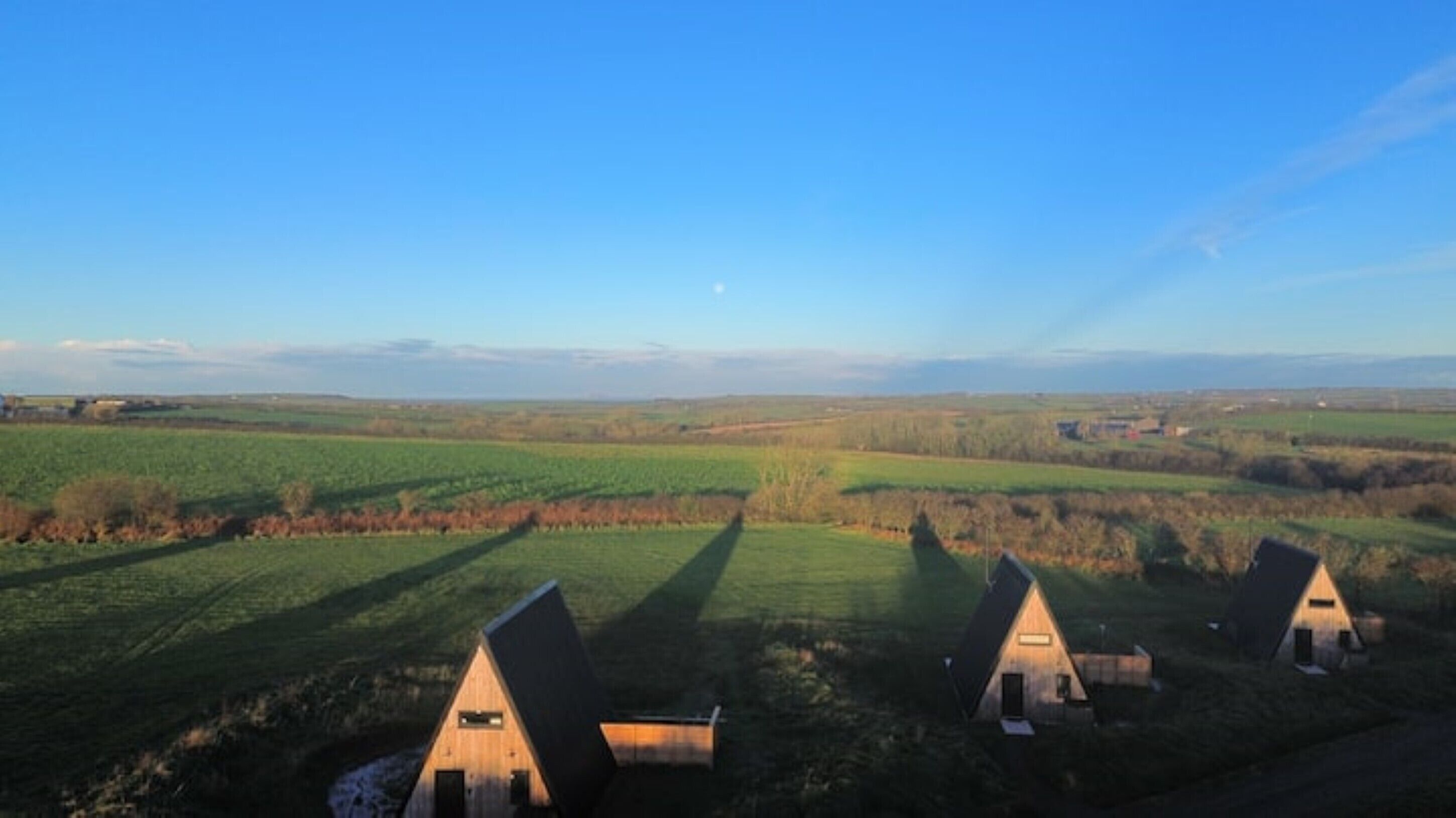 A rural landscape with three triangular-roofed cabins, a clear sky, and expansive fields.