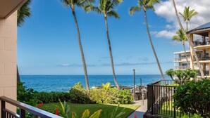 A coastal view with palm trees, a clear sky, and a building with balconies overlooking the ocean. - Kona Makai 1-103 Nearly Oceanfront, Ground Floor, No Stairs, Special Location (Kailua Kona)