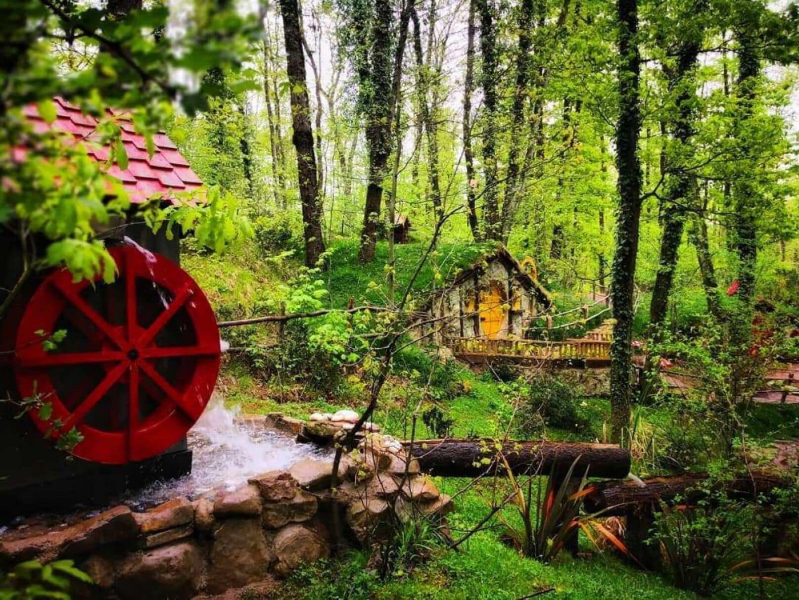A rustic wooden house surrounded by a lush forest, with a large red water wheel in the foreground.