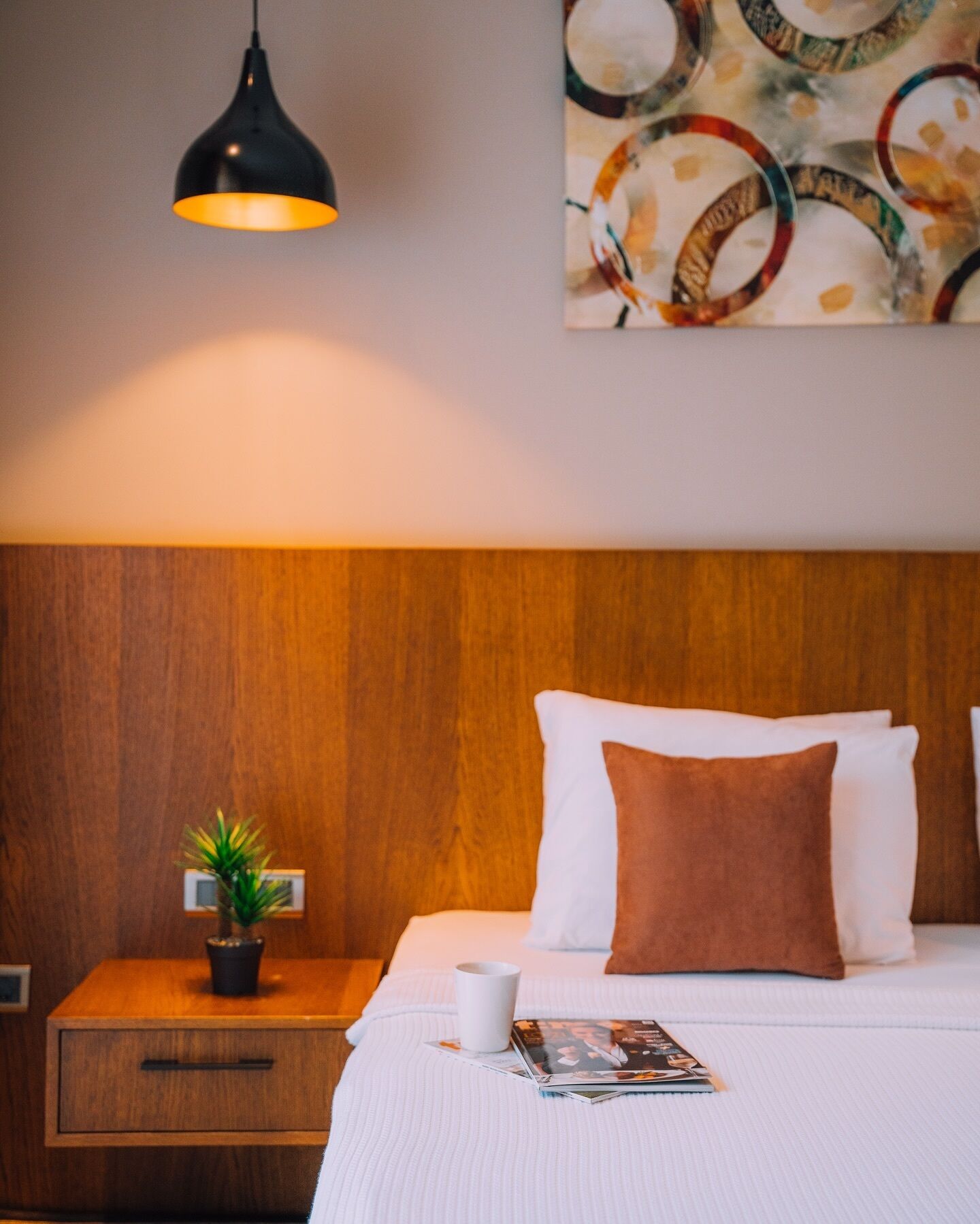 A neatly made bed with white and brown pillows, a bedside table with a plant, a magazine, and a black pendant light.