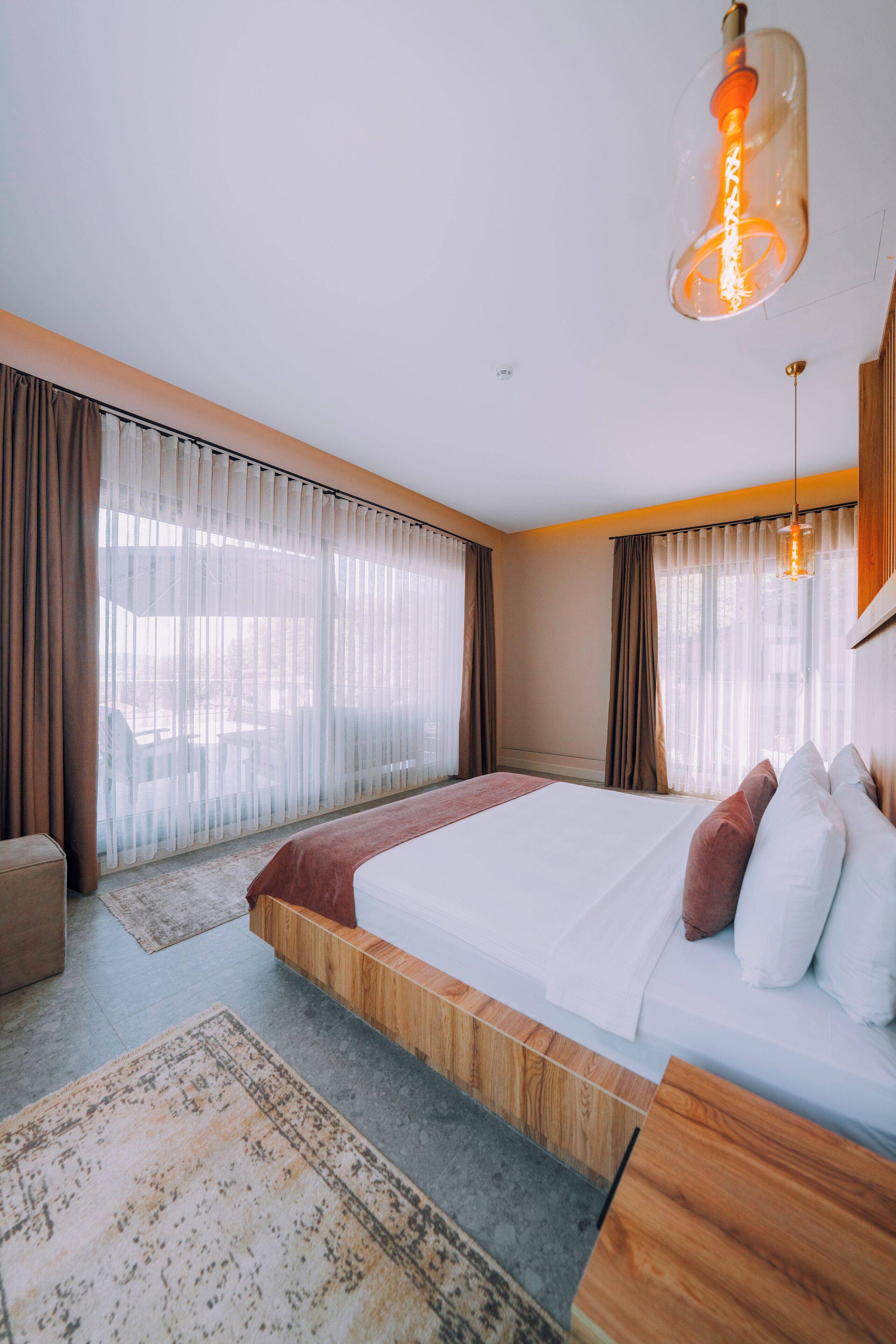 A modern hotel room with a large bed, wooden headboard, and a view of the outdoors through sheer curtains.