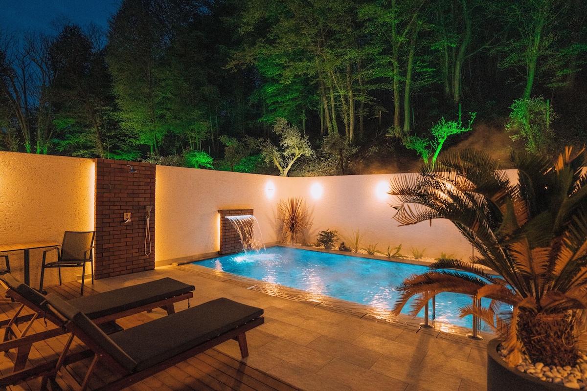 An outdoor swimming pool with a wooden deck, lounge chairs, and a brick wall.