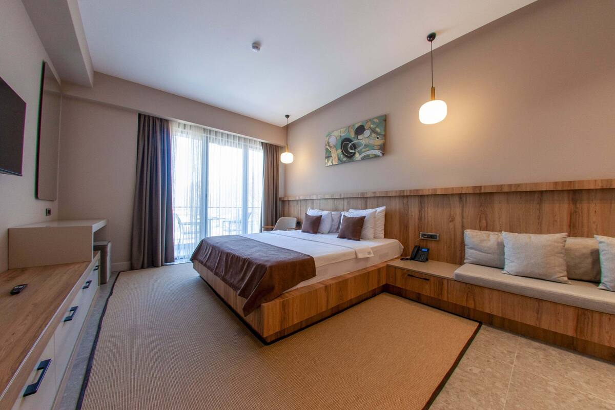 A modern hotel room with a large bed, bedside tables, a wooden headboard, a seating area, and a large window with curtains.