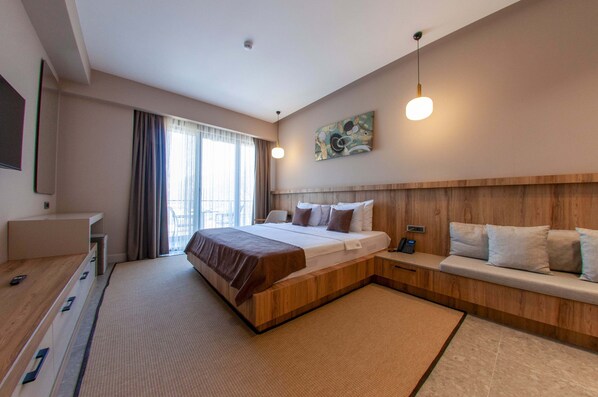 Une chambre d’hôtel moderne dotée d’un grand lit, de tables de chevet, d’une tête de lit en bois, d’un coin salon et d’une grande fenêtre avec des rideaux.