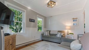 Un salón acogedor con un sofá gris, un sillón blanco y un aparador de madera. Hay dos ventanas con marcos blancos, un televisor de pantalla plana montado en la pared y una lámpara decorativa sobre una mesita.