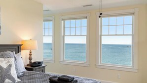 A bedroom with a bed, a bedside lamp, and three windows offering a view of the sea. - Overlook Beach House #4- York Maine Beach Rental (York)