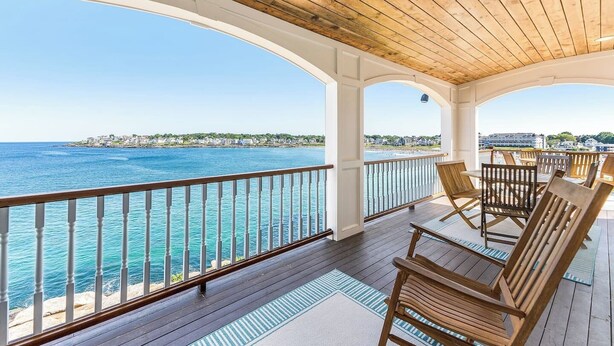 A covered outdoor deck with wooden chairs and a railing overlooking a body of water. - Overlook Beach House #4- York Maine Beach Rental (York)