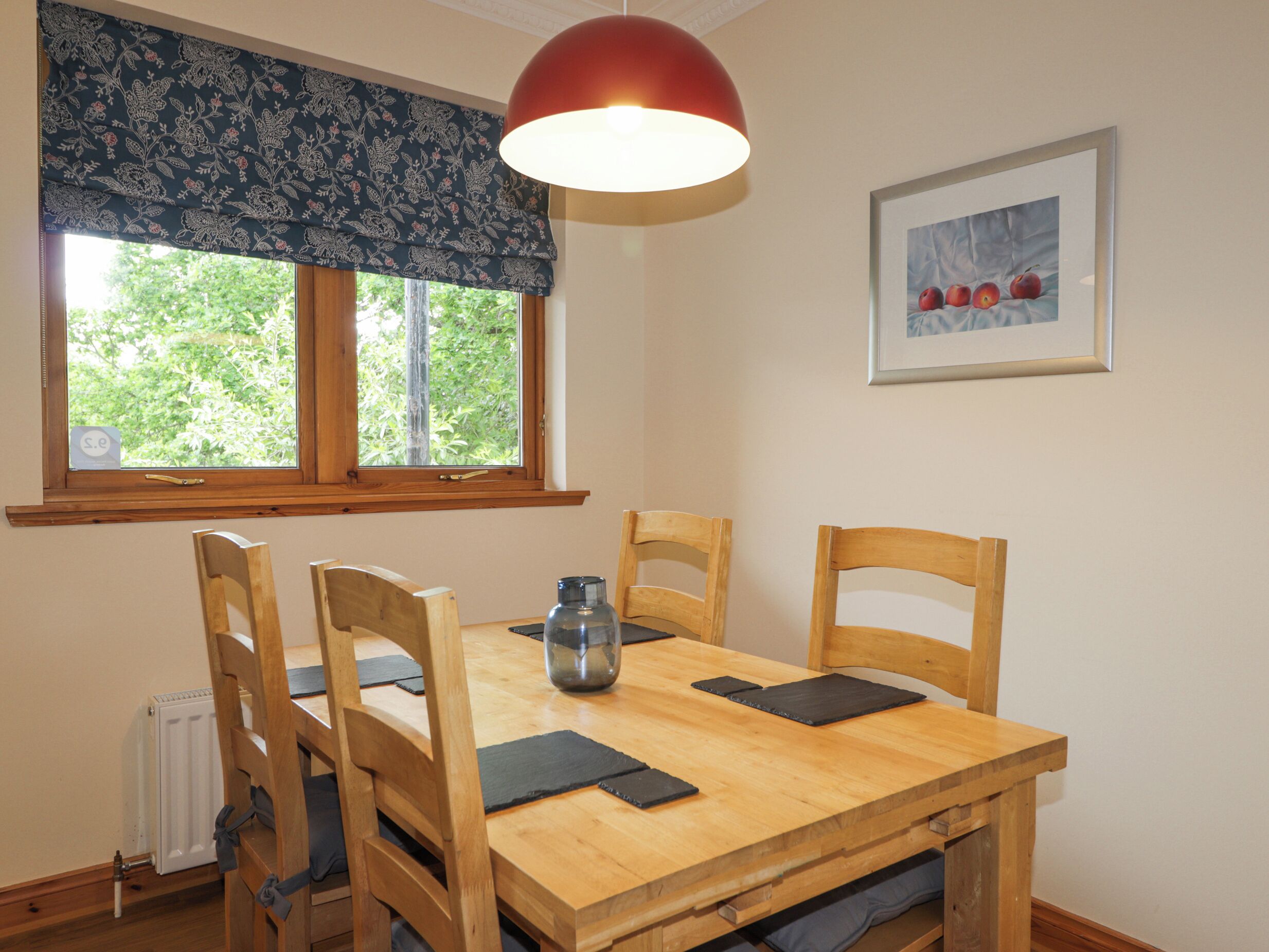 Une table à manger en bois, accompagnée de quatre chaises, un luminaire suspendu rouge, une fenêtre ornée d’une lambrequin à motifs floraux et un tableau encadré accroché au mur.