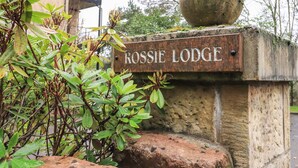 Ein rustikales Steinschild mit der Aufschrift "ROSSIE LODGE", angebracht an einer verwitterten Steinmauer, umgeben von Grünpflanzen und Steinen.