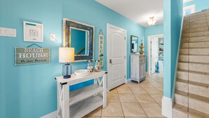A hallway with a blue wall, a white door, a mirror, and a sign that reads "BEACH HOUSE No shoes Required".