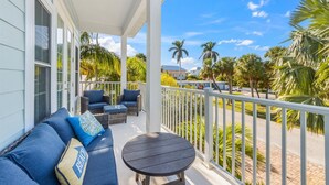 A covered outdoor seating area with a sofa and chairs, a round table, and a view of palm trees and a street.