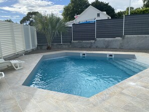 Une piscine avec une terrasse carrelée, un palmier et un bâtiment blanc en arrière-plan.