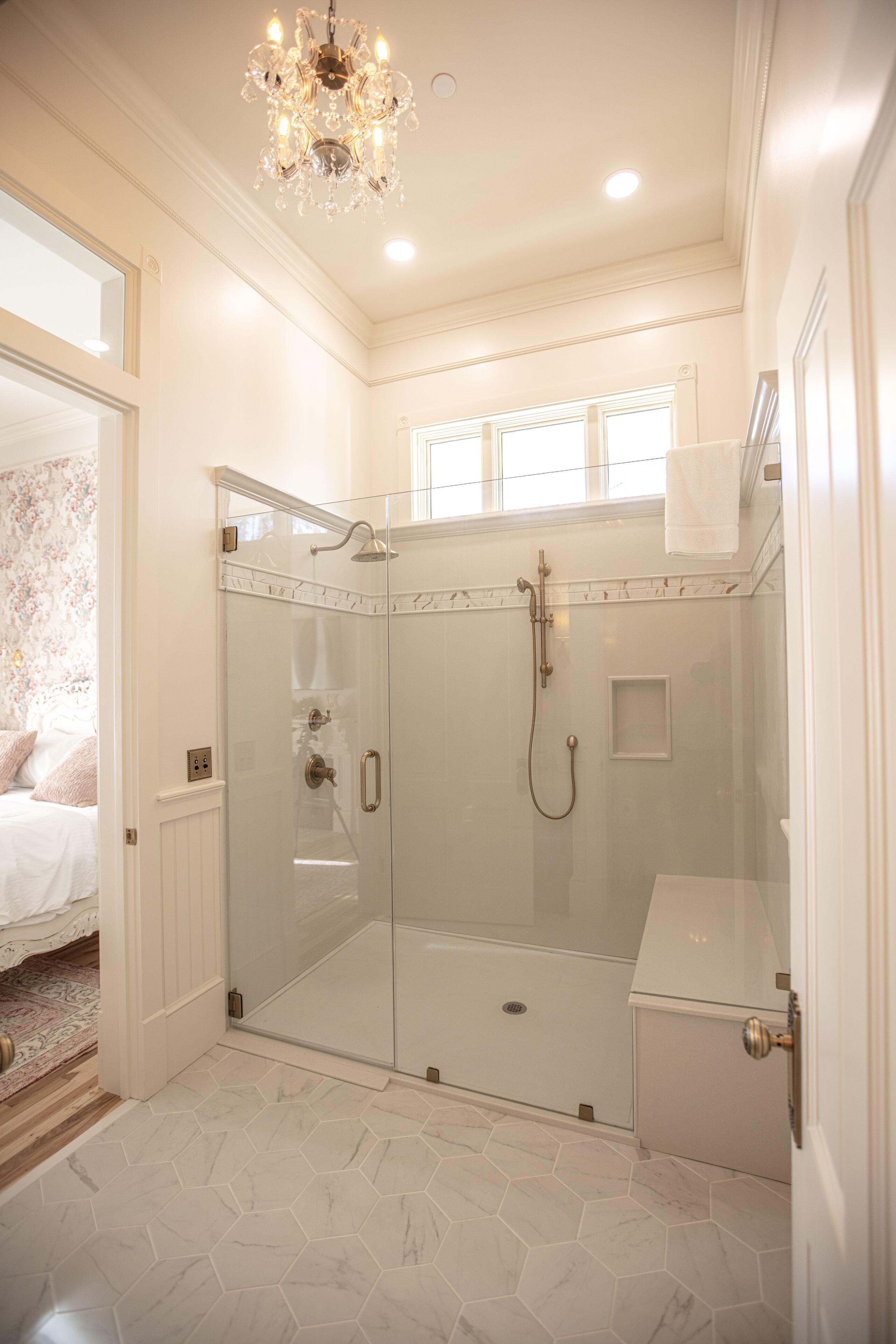 A modern bathroom with a glass-enclosed shower, a chandelier, and a view of a bedroom with floral wallpaper.