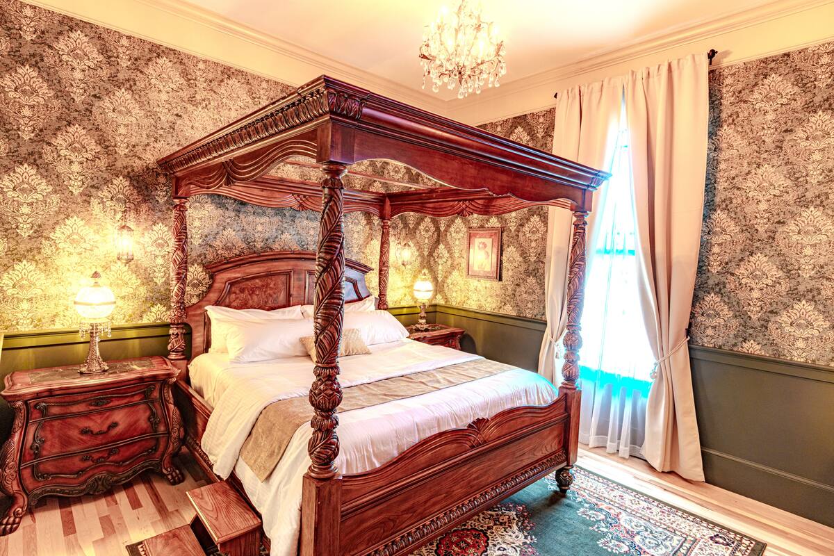 A four-poster bed with ornate wooden carvings, a chandelier, and a patterned wallpaper.