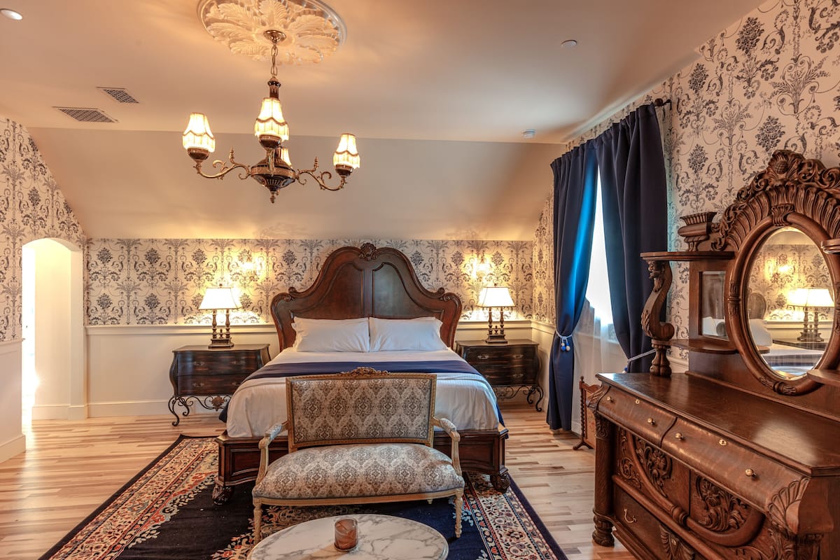 A bedroom with a large bed, two bedside tables with lamps, a dresser with a mirror, a chair, and a patterned rug.