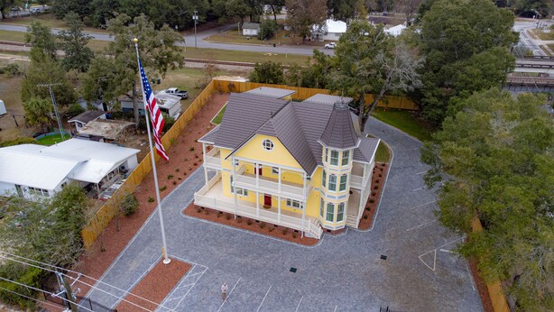 A two-story building with a yellow facade and a grey roof, surrounded by a fenced yard and trees.