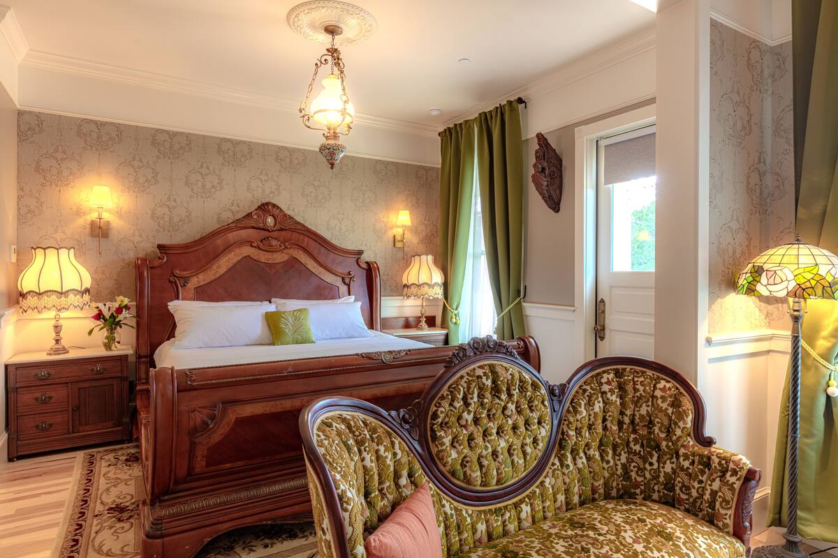 A bedroom with a wooden four-poster bed, two upholstered chairs, a nightstand with lamps, and a window with curtains.