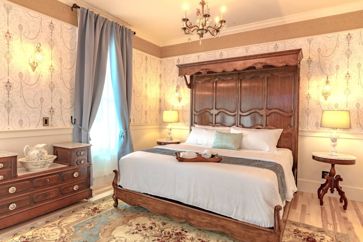 A classic bedroom with a wooden bed, two bedside tables with lamps, a dresser, and patterned wallpaper.