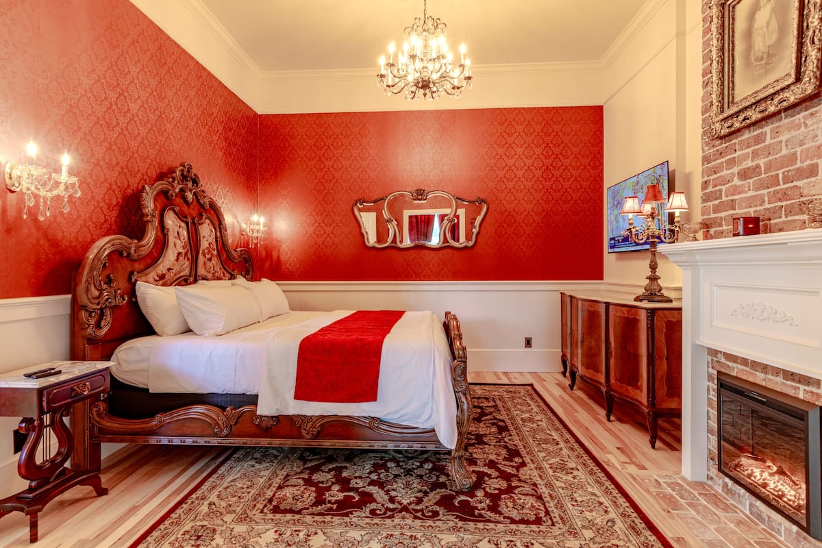 A bedroom with a large ornate bed, a red accent wall, a chandelier, a fireplace, and a red rug.