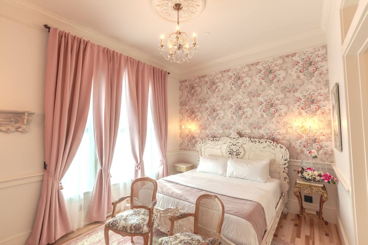 A bedroom with a large bed, two chairs, a nightstand with a lamp, and a floral wallpaper.