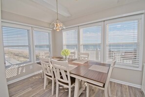 Un coin repas avec une table en bois et des chaises blanches, de grandes fenêtres équipées de stores et un luminaire suspendu.