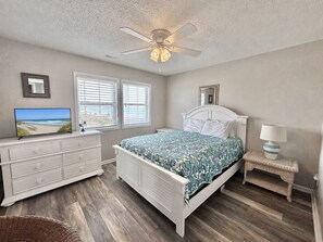 Une chambre à coucher comprenant un lit, une commode, un téléviseur, un ventilateur de plafond et une fenêtre avec des stores.
