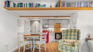 A compact living space with a dining area, kitchen, and a bookshelf filled with books. - The Cwtch Apartment - Sea Views, Walk to Beach (Penally)
