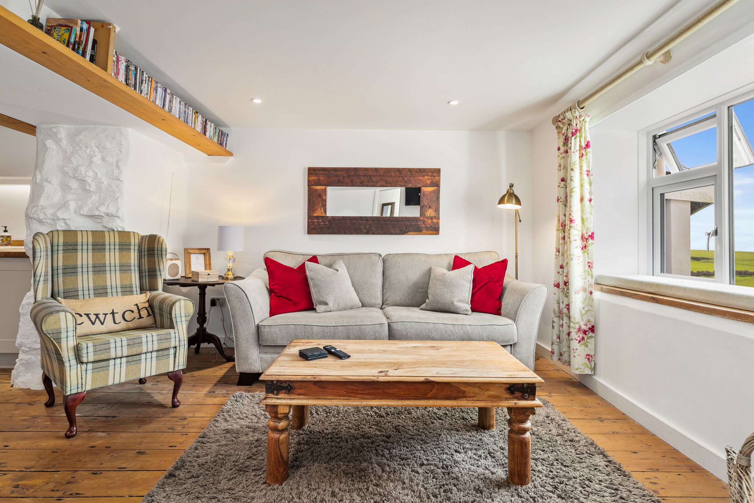 A cozy living room with a plaid armchair, a grey sofa with red cushions, a wooden coffee table, and a bookshelf filled with books.