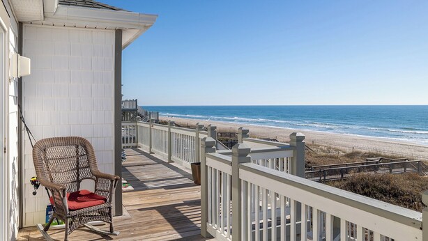 Terraza de madera con una silla de mimbre y un cojín rojo, con vistas a una playa que cuenta con barandilla de madera y cielo despejado.