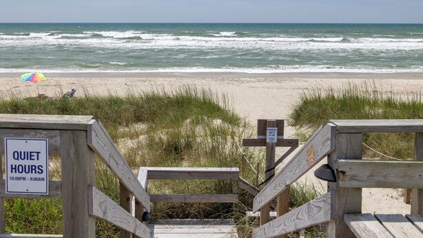 Blick auf einen Strand mit einer Holztreppe, die zum Sand führt, einem Schild mit der Aufschrift Ruhezeiten und einem Strandsonnenschirm.