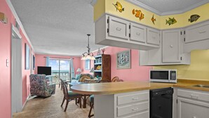A kitchen with white cabinets, a wooden countertop, and a dishwasher. There is a dining area with a table and chairs, and a view of the outdoors through large windows. - Point Emerald A-205, Oceanfront Condo in Emerald Isle, NC! (Emerald Isle)