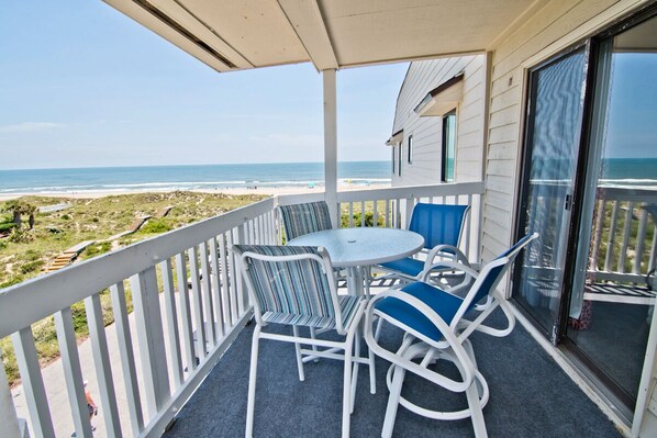 A balcony with a view of the beach, white outdoor furniture, and a glass door.