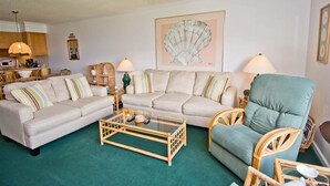 A living room with a beige sofa, a green armchair, a glass coffee table, and a large shell artwork on the wall.