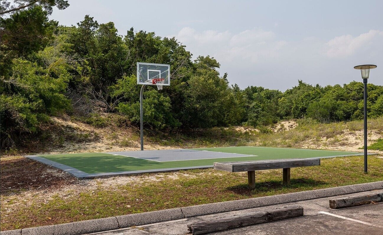 A basketball court with a hoop and a bench, surrounded by trees and a lamp post.