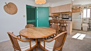 Ein Essbereich mit einem runden Holztisch und Korbstühlen, einer grünen Tür sowie einer Küche mit Holzschränken und einem Kühlschrank.