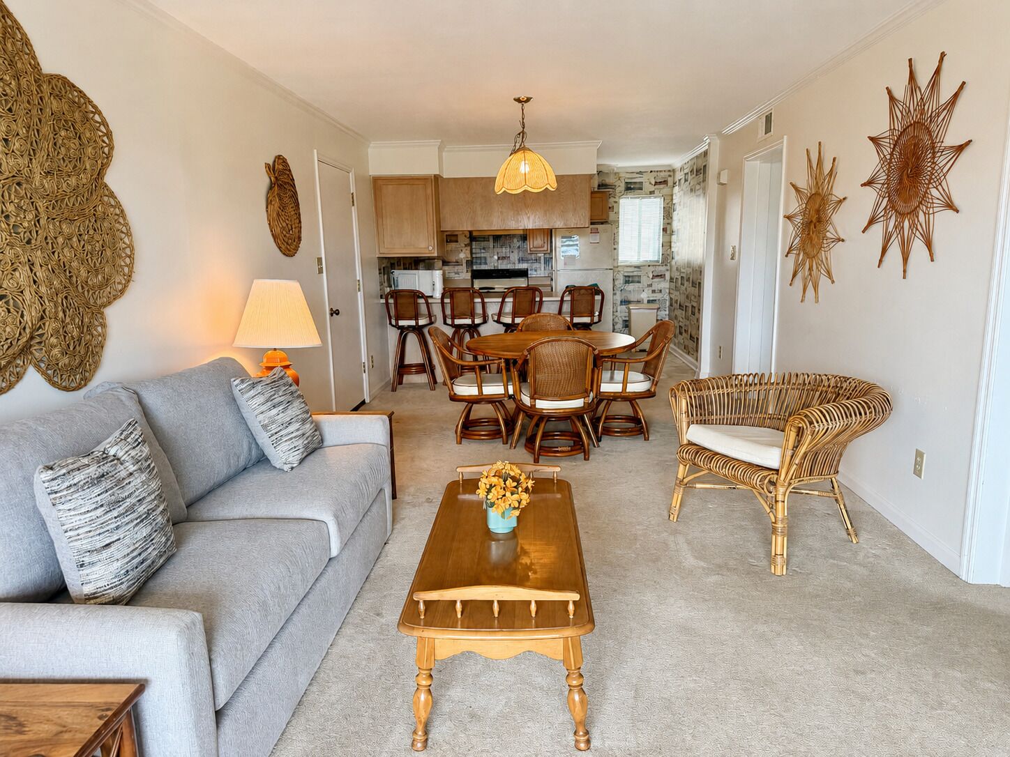 A living room with a sofa, coffee table, and wicker chairs. There is a dining area with a table and chairs, and a kitchen in the background.