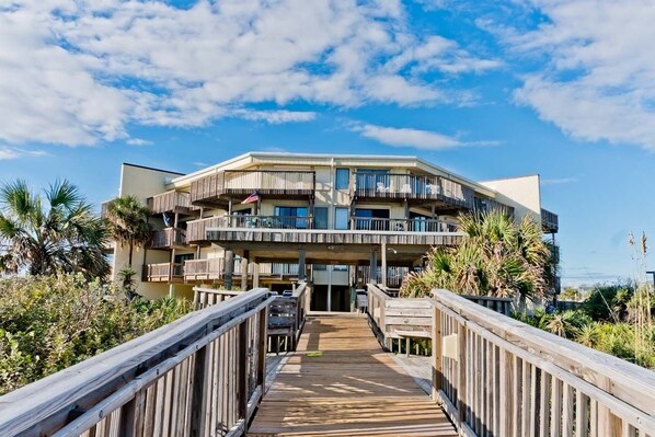 A beachfront hotel with a wooden boardwalk leading to the entrance, multiple balconies, and a clear view of the ocean.