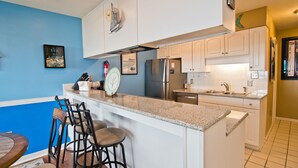 A modern kitchen with white cabinets, a granite countertop, and stainless steel appliances. There is a bar area with stools and a clock on the wall. - Queens Court 1204, Oceanfront Condo in Emerald Isle, NC! (Emerald Isle)