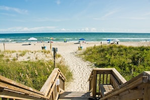Een houten looppad dat naar een zandstrand leidt, met parasols en mensen.