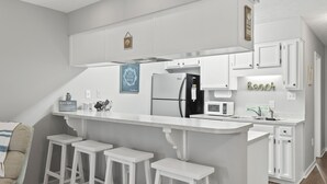 Eine moderne Küche mit weißen Schränken, einer zentralen Kochinsel mit Barhocker, einem Kühlschrank, einer Mikrowelle und einem Spülbecken.