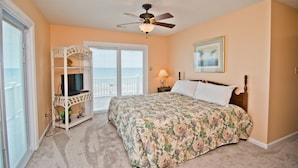 A bedroom with a bed, a ceiling fan, a TV on a white stand, a window with a view, and a framed picture on the wall. - Red Roof Cottage East, Oceanfront Duplex in Emerald Isle, NC! (Emerald Isle)