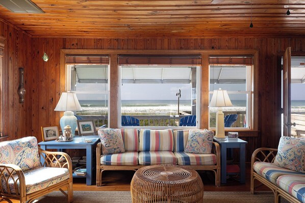 A cozy room with a wooden ceiling, a striped sofa, wicker chairs, and large windows overlooking the beach.