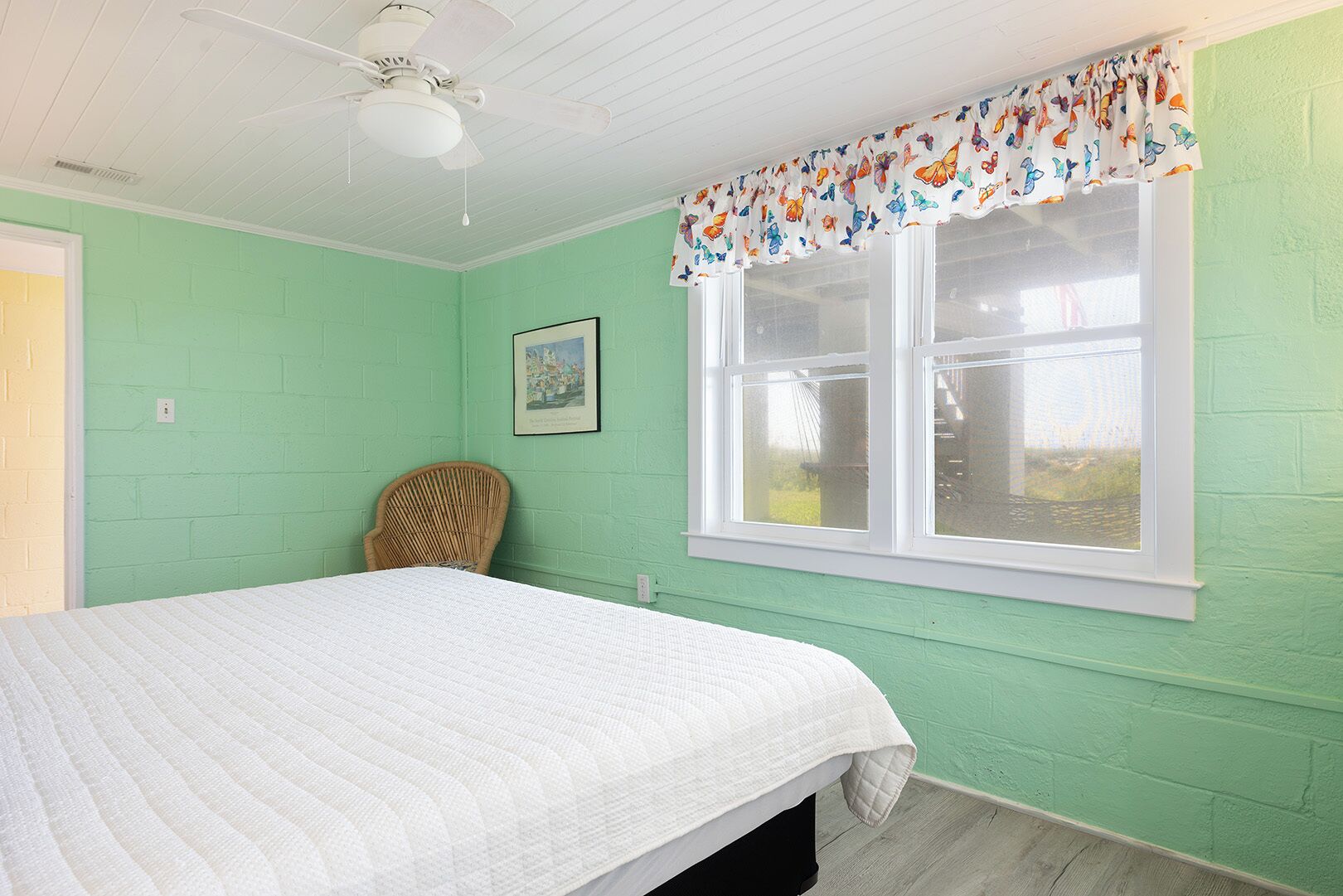 A room with a bed, a window with a butterfly-patterned valance, a ceiling fan, and a wicker chair.